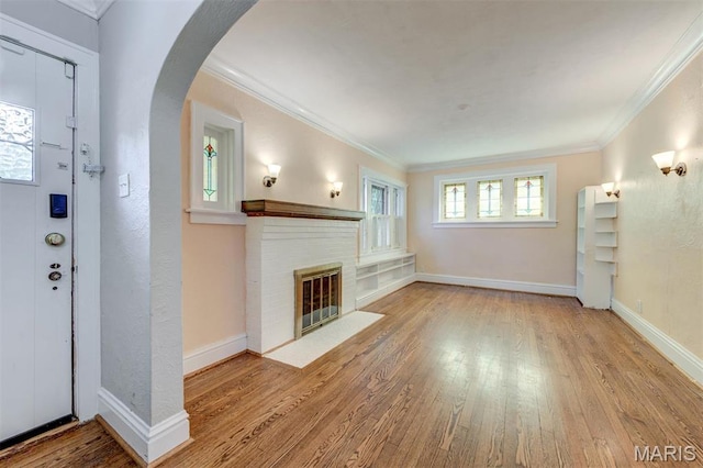 unfurnished living room with wood finished floors, ornamental molding, arched walkways, a brick fireplace, and baseboards