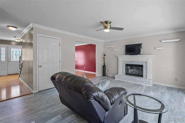 living room featuring ceiling fan, wood finished floors, crown molding, baseboards, and a brick fireplace