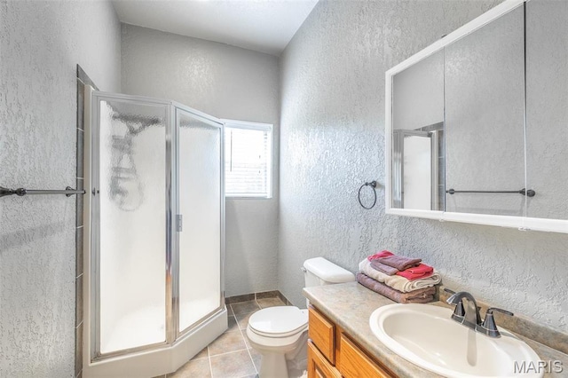 bathroom with a textured wall, a shower stall, toilet, vanity, and tile patterned flooring
