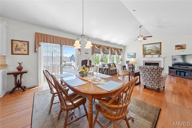 dining space with a ceiling fan, light wood finished floors, a fireplace, vaulted ceiling, and a chandelier