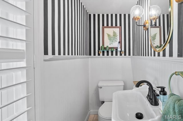 half bath with a sink, a chandelier, wallpapered walls, a wainscoted wall, and toilet