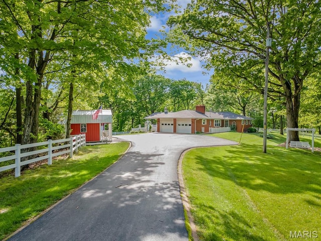 view of front of property featuring asphalt driveway, a garage, a chimney, view of wooded area, and an outbuilding