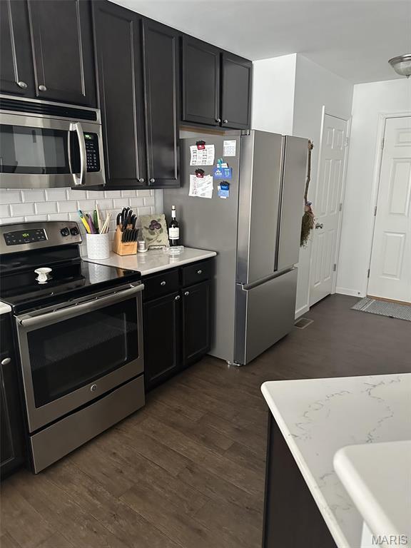 1805 Hyland Green Dr, Ofallon, MO 63366 - kitchen with appliances with stainless steel finishes, dark cabinetry, decorative backsplash, dark wood-type flooring, and light stone counters