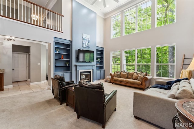 living area featuring a towering ceiling, a glass covered fireplace, a ceiling fan, light carpet, and baseboards