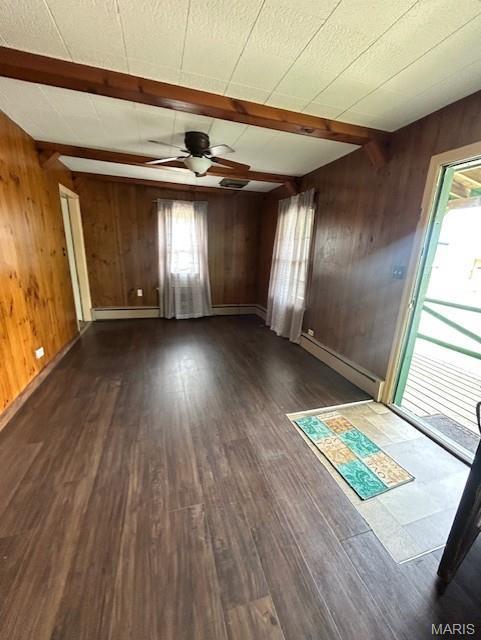 unfurnished living room featuring beam ceiling, a ceiling fan, wooden walls, plenty of natural light, and dark wood-type flooring