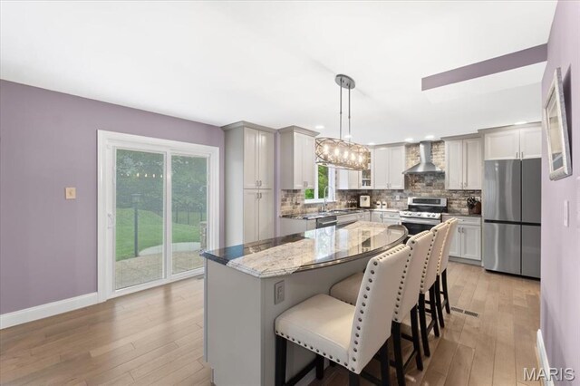kitchen featuring appliances with stainless steel finishes, wall chimney range hood, decorative backsplash, light wood-style flooring, and a kitchen breakfast bar