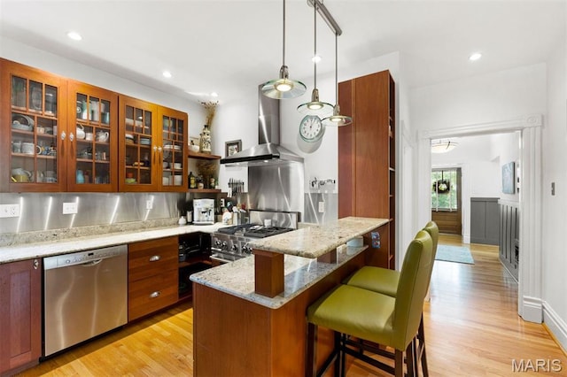 kitchen featuring appliances with stainless steel finishes, wall chimney exhaust hood, a breakfast bar area, light wood-style floors, and glass insert cabinets