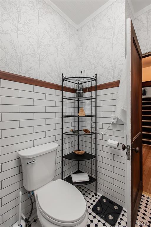 bathroom with crown molding, wallpapered walls, toilet, tiled shower, and a wainscoted wall