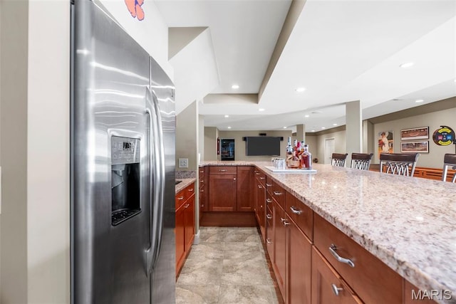 kitchen featuring stainless steel refrigerator with ice dispenser, light stone countertops, recessed lighting, brown cabinetry, and a peninsula