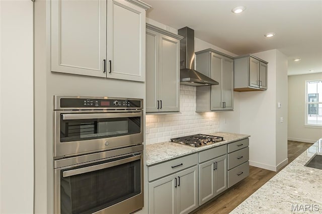 kitchen with appliances with stainless steel finishes, wall chimney range hood, gray cabinets, tasteful backsplash, and dark wood-type flooring