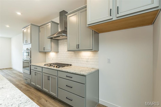 kitchen featuring gray cabinets, wall chimney range hood, stainless steel appliances, baseboards, and decorative backsplash