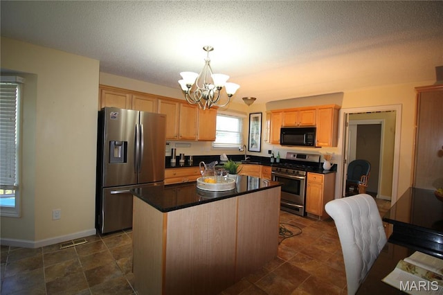 kitchen featuring stainless steel appliances, a chandelier, light brown cabinetry, dark countertops, and a kitchen island