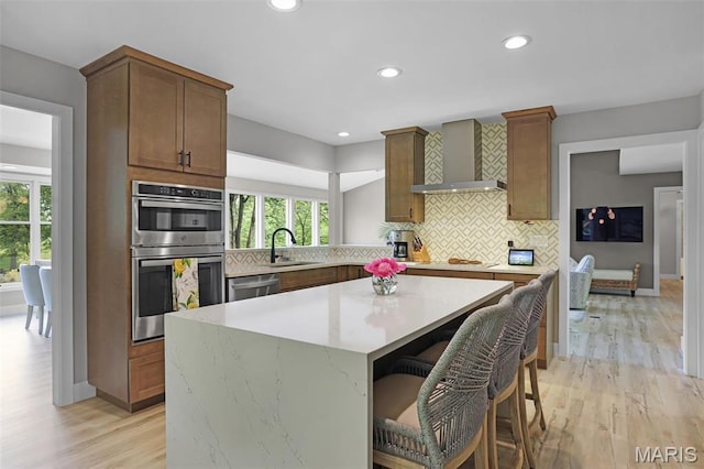 kitchen featuring wall chimney range hood, stainless steel appliances, a kitchen island, tasteful backsplash, and light wood-style floors