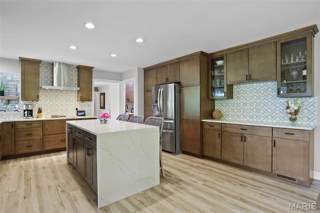 kitchen with stainless steel refrigerator with ice dispenser, wall chimney range hood, tasteful backsplash, light wood-style flooring, and recessed lighting