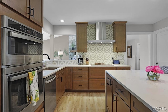 kitchen featuring wall chimney exhaust hood, stainless steel appliances, a sink, light wood-style floors, and light stone counters