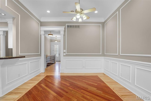 unfurnished dining area featuring a decorative wall, a ceiling fan, ornamental molding, light wood-style flooring, and ornate columns