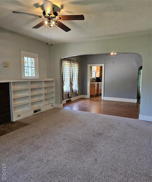 unfurnished living room featuring arched walkways, dark colored carpet, a ceiling fan, a textured ceiling, and baseboards