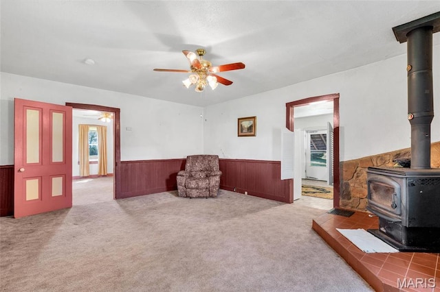 living area featuring a wood stove, a ceiling fan, wainscoting, and carpet floors