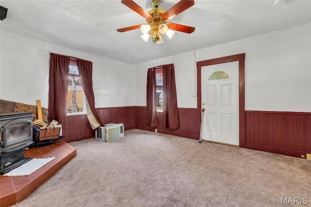 unfurnished living room featuring a wood stove, a wainscoted wall, ceiling fan, wood walls, and carpet floors