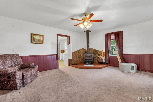 living room with a wood stove, a wainscoted wall, ceiling fan, carpet, and wooden walls