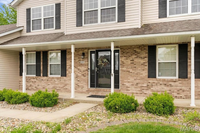 doorway to property featuring covered porch, brick siding, and roof with shingles