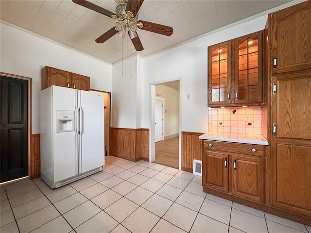 kitchen featuring white fridge with ice dispenser, wainscoting, tile countertops, ceiling fan, and brown cabinets