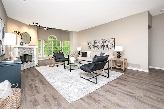 living area featuring ceiling fan, wood finished floors, baseboards, a stone fireplace, and high vaulted ceiling