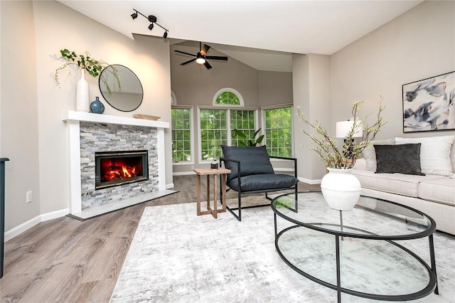 living area with a stone fireplace, a ceiling fan, wood finished floors, baseboards, and high vaulted ceiling
