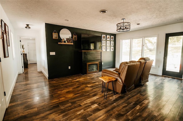 living area featuring a textured ceiling, dark wood-style flooring, a glass covered fireplace, a chandelier, and baseboards