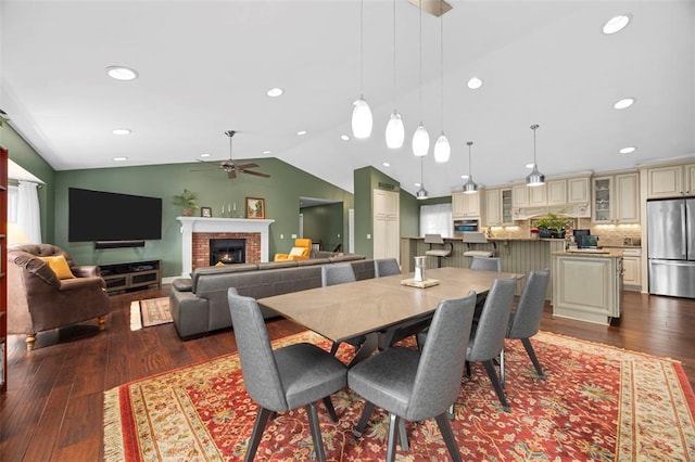 dining room with a ceiling fan, lofted ceiling, dark wood-style floors, a brick fireplace, and recessed lighting
