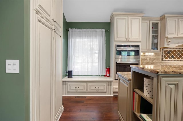 kitchen featuring stainless steel double oven, cream cabinetry, decorative backsplash, dark wood-style floors, and light stone countertops