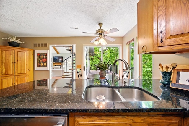 kitchen with a sink, dishwasher, brown cabinetry, a textured ceiling, and dark stone counters