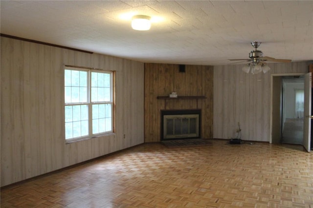 unfurnished living room with baseboards, wood walls, a large fireplace, a ceiling fan, and parquet floors