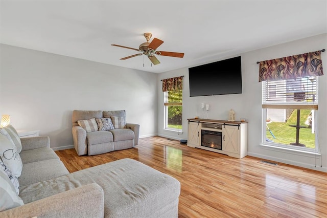 living area featuring light wood-type flooring, a glass covered fireplace, baseboards, and ceiling fan