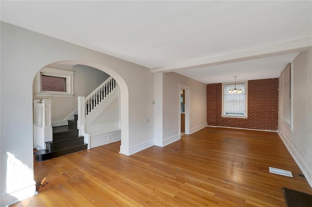 unfurnished living room with light wood-style flooring, stairs, brick wall, a chandelier, and baseboards