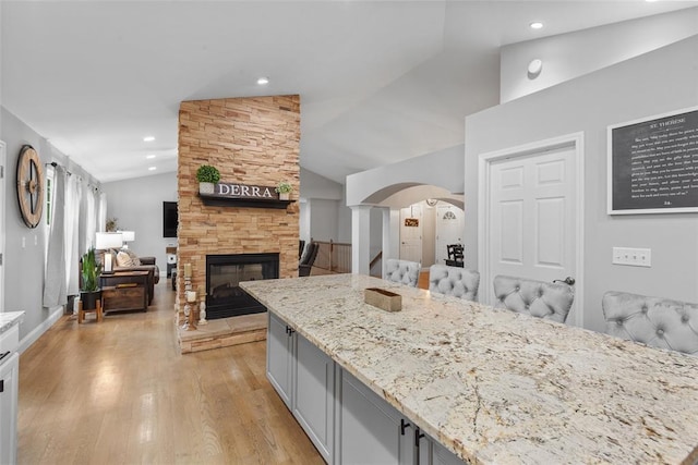 kitchen with arched walkways, vaulted ceiling, light stone counters, a fireplace, and light wood finished floors