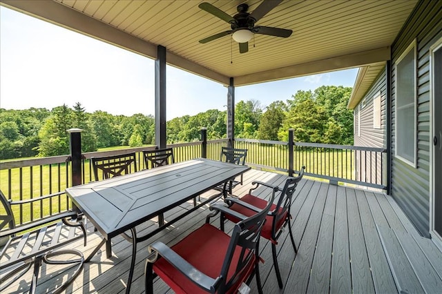 deck featuring outdoor dining area, ceiling fan, a yard, and view of wooded area