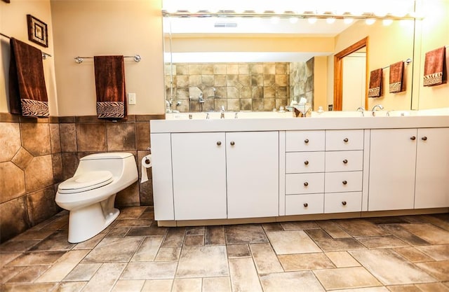 bathroom featuring toilet, double vanity, stone finish flooring, tile walls, and a wainscoted wall