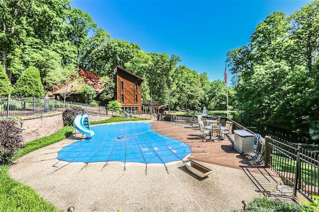 view of pool with a water slide, fence, a wooden deck, view of scattered trees, and a patio area