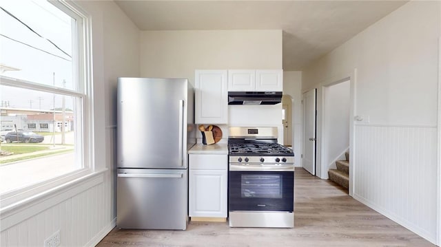 kitchen with stainless steel appliances, under cabinet range hood, healthy amount of natural light, white cabinetry, and wainscoting