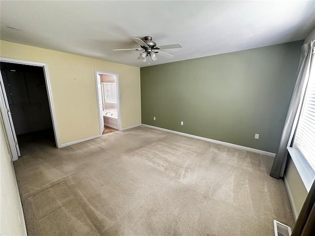unfurnished bedroom with light carpet, baseboards, a walk in closet, ensuite bathroom, and ceiling fan