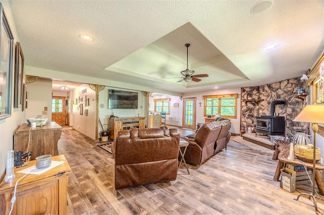 living room featuring a ceiling fan, a wood stove, light wood-style floors, a textured ceiling, and a raised ceiling