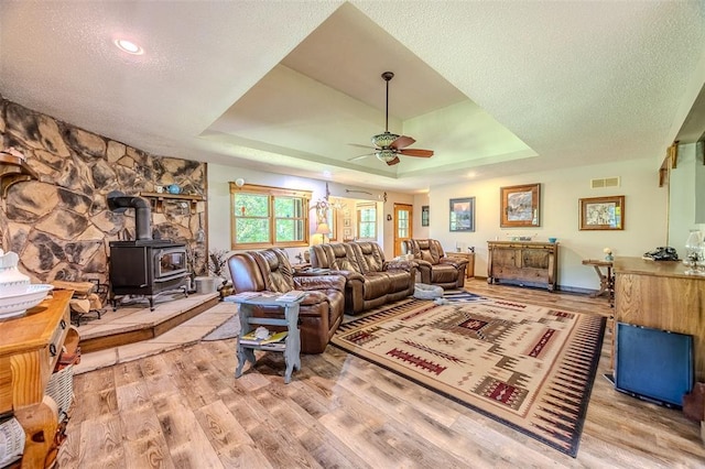 living room featuring ceiling fan, a wood stove, a tray ceiling, wood finished floors, and a textured ceiling