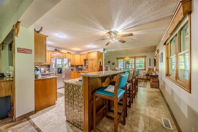 kitchen featuring stainless steel fridge, a textured ceiling, a ceiling fan, light stone countertops, and a kitchen bar