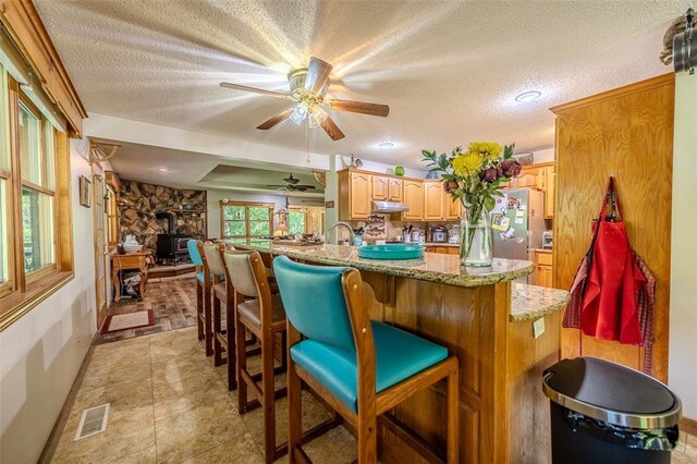 kitchen featuring a wood stove, a textured ceiling, a ceiling fan, under cabinet range hood, and light stone counters