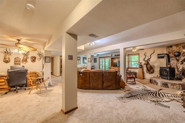 living area with ceiling fan, a wood stove, light carpet, a desk, and a textured ceiling