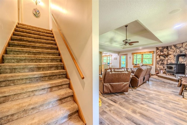 stairway featuring a tray ceiling, a wood stove, a ceiling fan, wood finished floors, and a textured ceiling