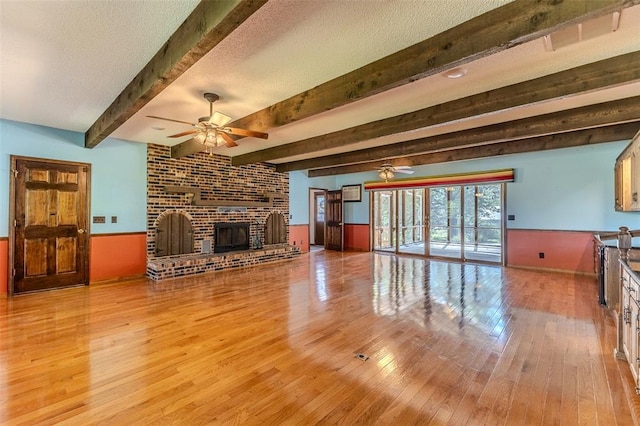 21509 Rockwell Dr, Lebanon, MO 65536 - unfurnished living room featuring a ceiling fan, wood finished floors, beamed ceiling, a brick fireplace, and a wainscoted wall