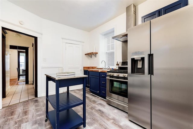 4219 Red Bud Ave, Saint Louis, MO 63115 - kitchen with appliances with stainless steel finishes, blue cabinetry, butcher block countertops, wall chimney range hood, and light wood-style flooring