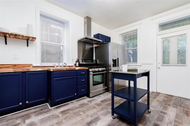 4219 Red Bud Ave, Saint Louis, MO 63115 - kitchen with blue cabinetry, appliances with stainless steel finishes, wooden counters, wall chimney exhaust hood, and a sink